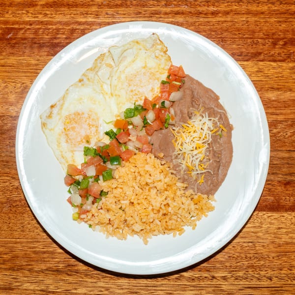 A white plate holds two fried eggs, pico de gallo, rice, and refried beans with shredded cheese.