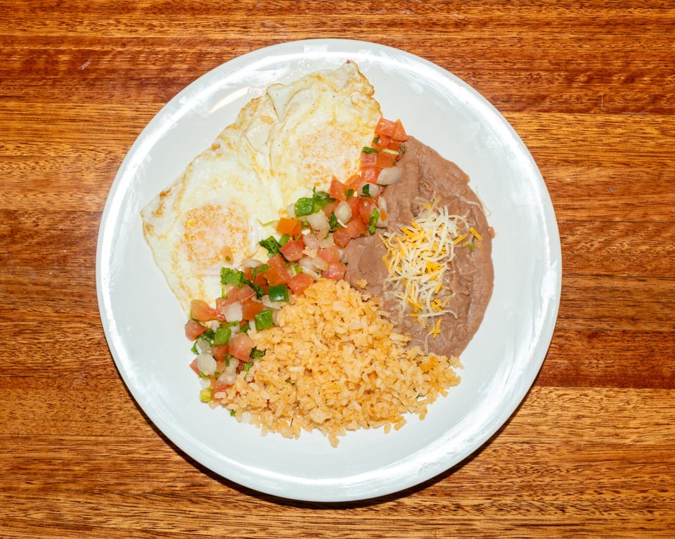 A white plate holds two fried eggs, pico de gallo, rice, and refried beans with shredded cheese.