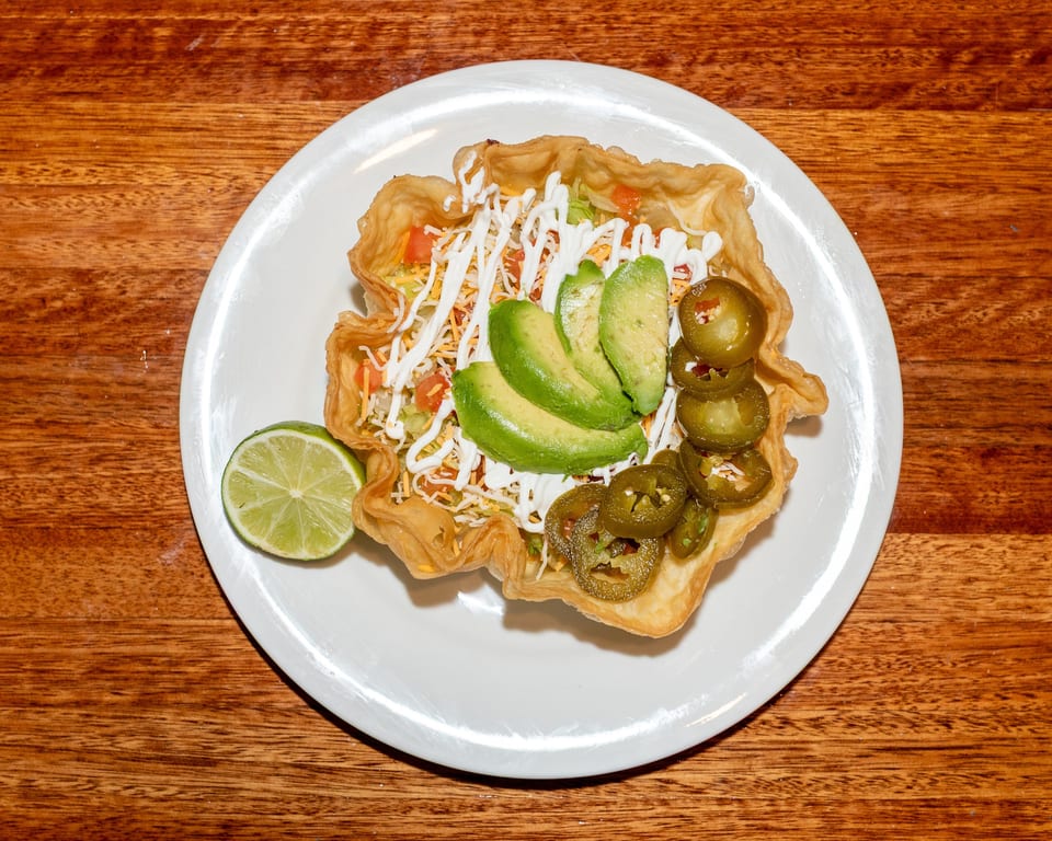 Taco salad in a crispy tortilla bowl with avocado, jalapeños, sour cream, and a lime wedge.