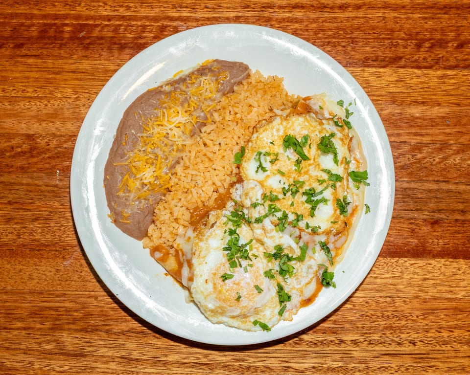 A white plate with huevos rancheros, refried beans topped with cheese, and Spanish rice.