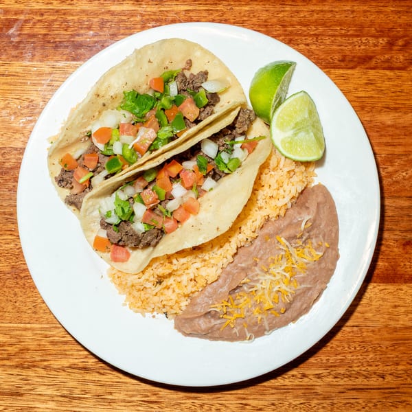 Two beef tacos with pico de gallo, rice, refried beans, and lime wedges on a white plate.