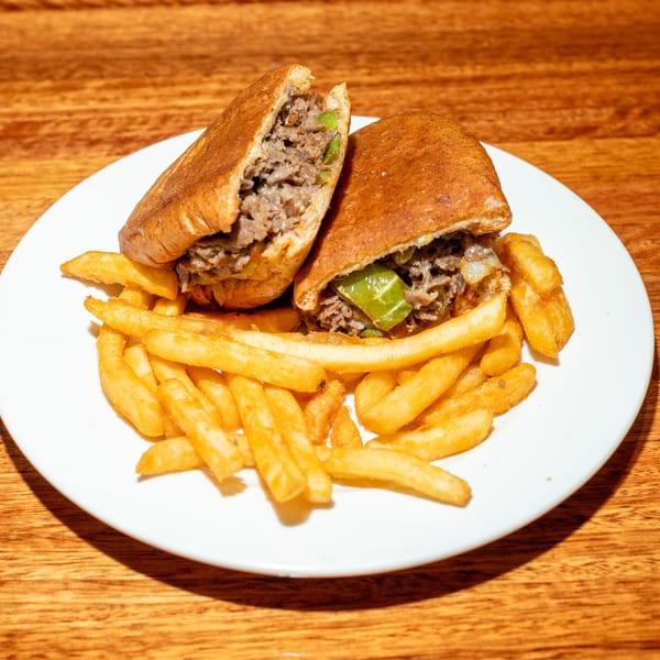 Cheesesteak sandwich cut in half, filled with meat, onions, and peppers, served with fries.