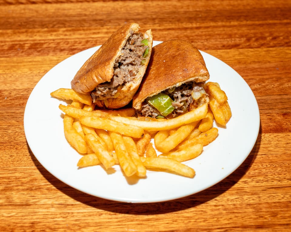 Cheesesteak sandwich cut in half, filled with meat, onions, and peppers, served with fries.