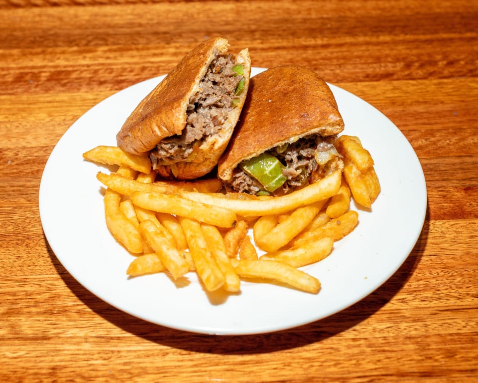 A cheesesteak sandwich, cut in half, with fries on a white plate on a wooden table.