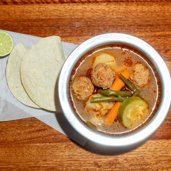 Albondigas soup with meatballs, vegetables, tortillas, and a lime wedge on a wooden table.