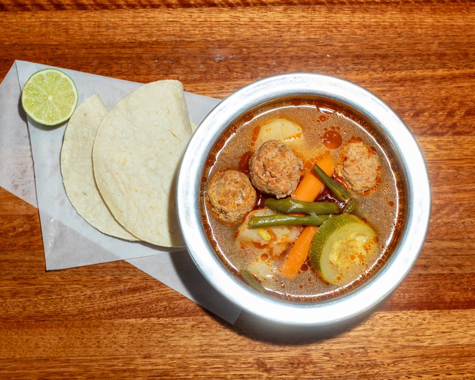 Albondigas soup with meatballs, vegetables, tortillas, and a lime wedge on a wooden table.