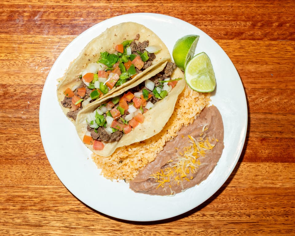 Two beef tacos with pico de gallo, rice, refried beans, and lime wedges on a white plate.